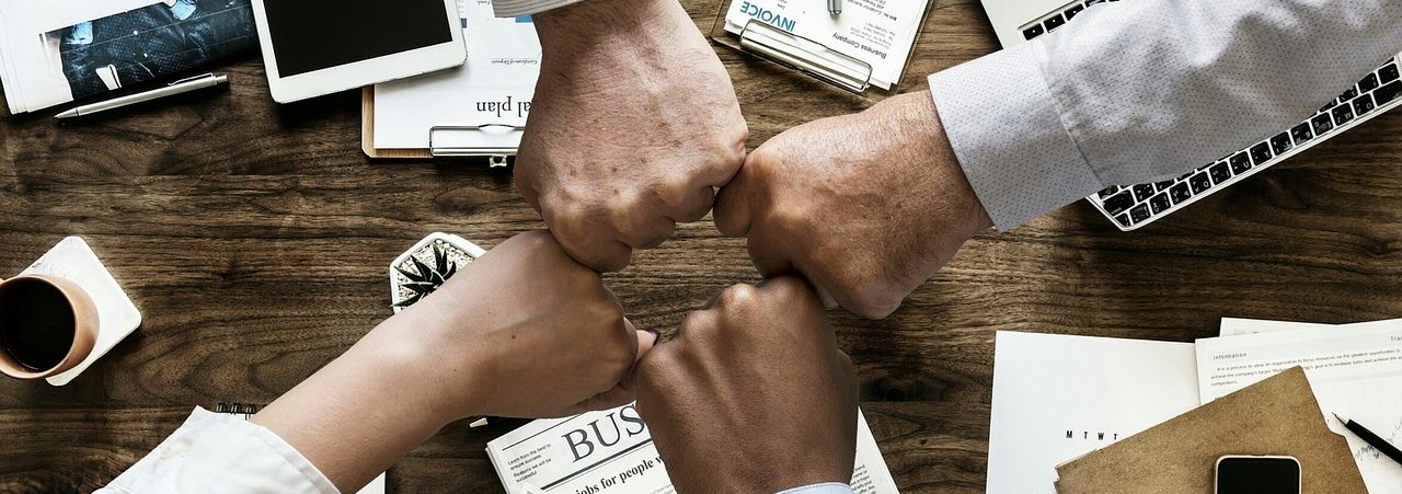 Four colleagues bumping fists over a desk of paperwork and laptops